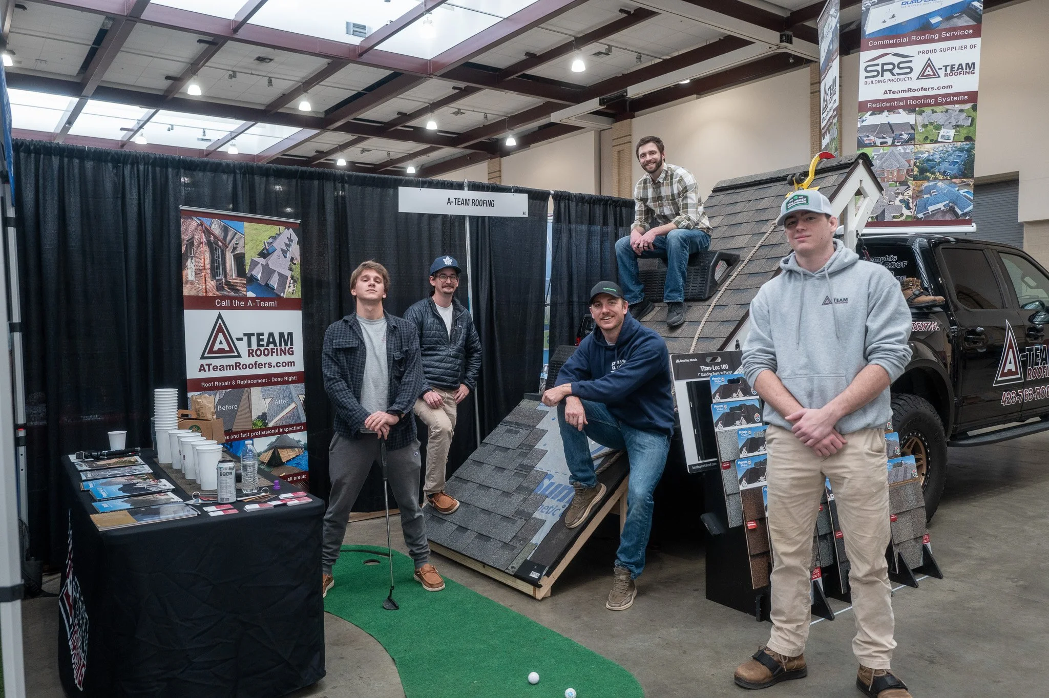 A group of five men at a roofing booth displaying roofing samples and information, with a small golf putting green and a roof display in an indoor exhibition hall.
