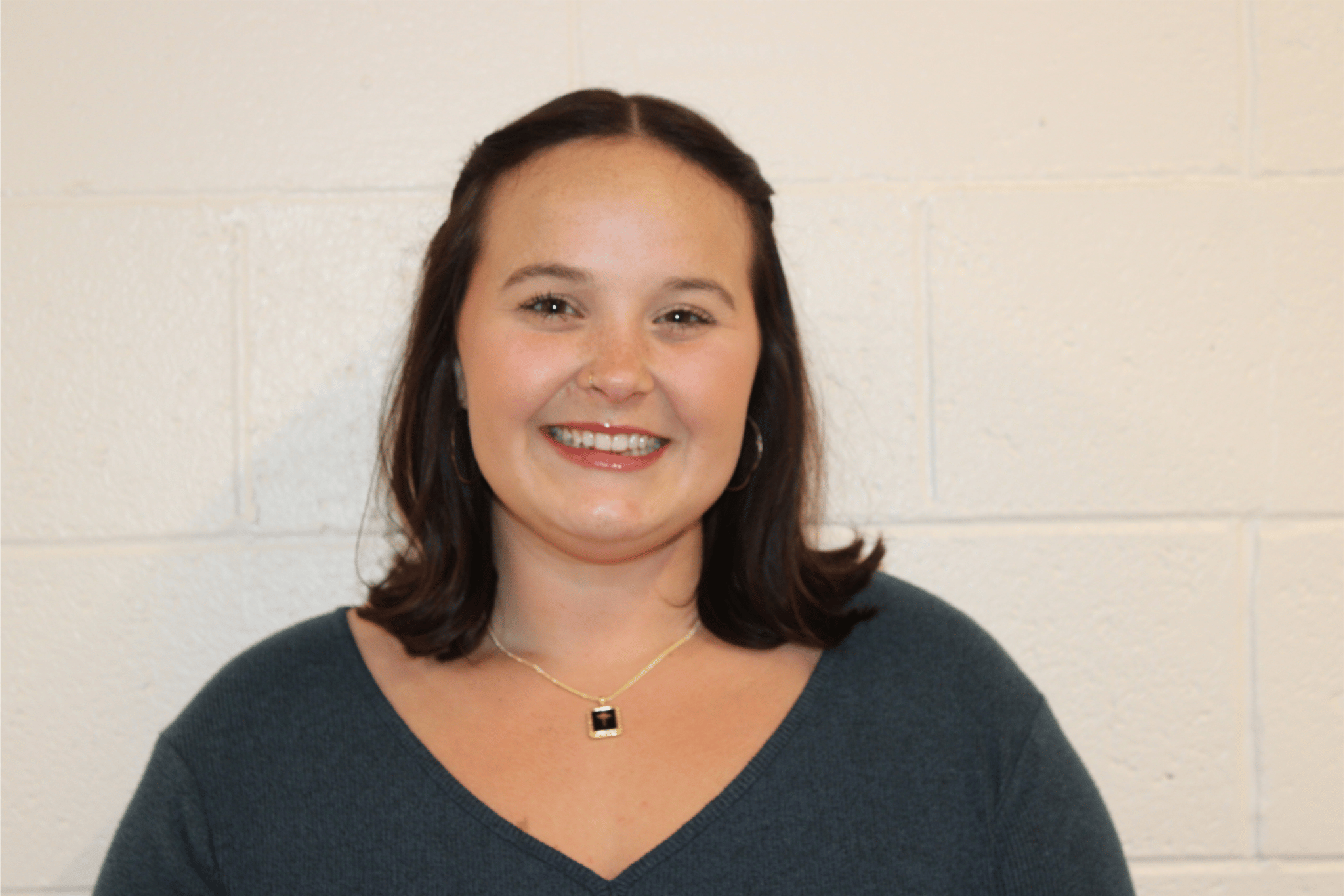 A woman with medium-length brown hair, smiling, wearing a navy sweater, a gold necklace with a pendant, and earrings, standing against a white brick wall.