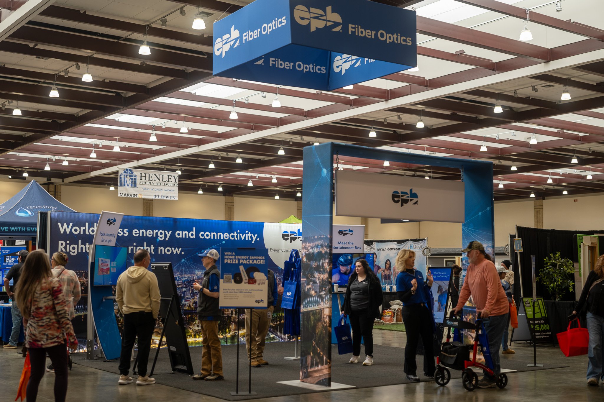 At a trade show, a blue booth displays 'EPB Fiber Optics' signage with a digital-themed backdrop. Attendees walk and browse different informational displays in a large, well-lit exhibition hall.