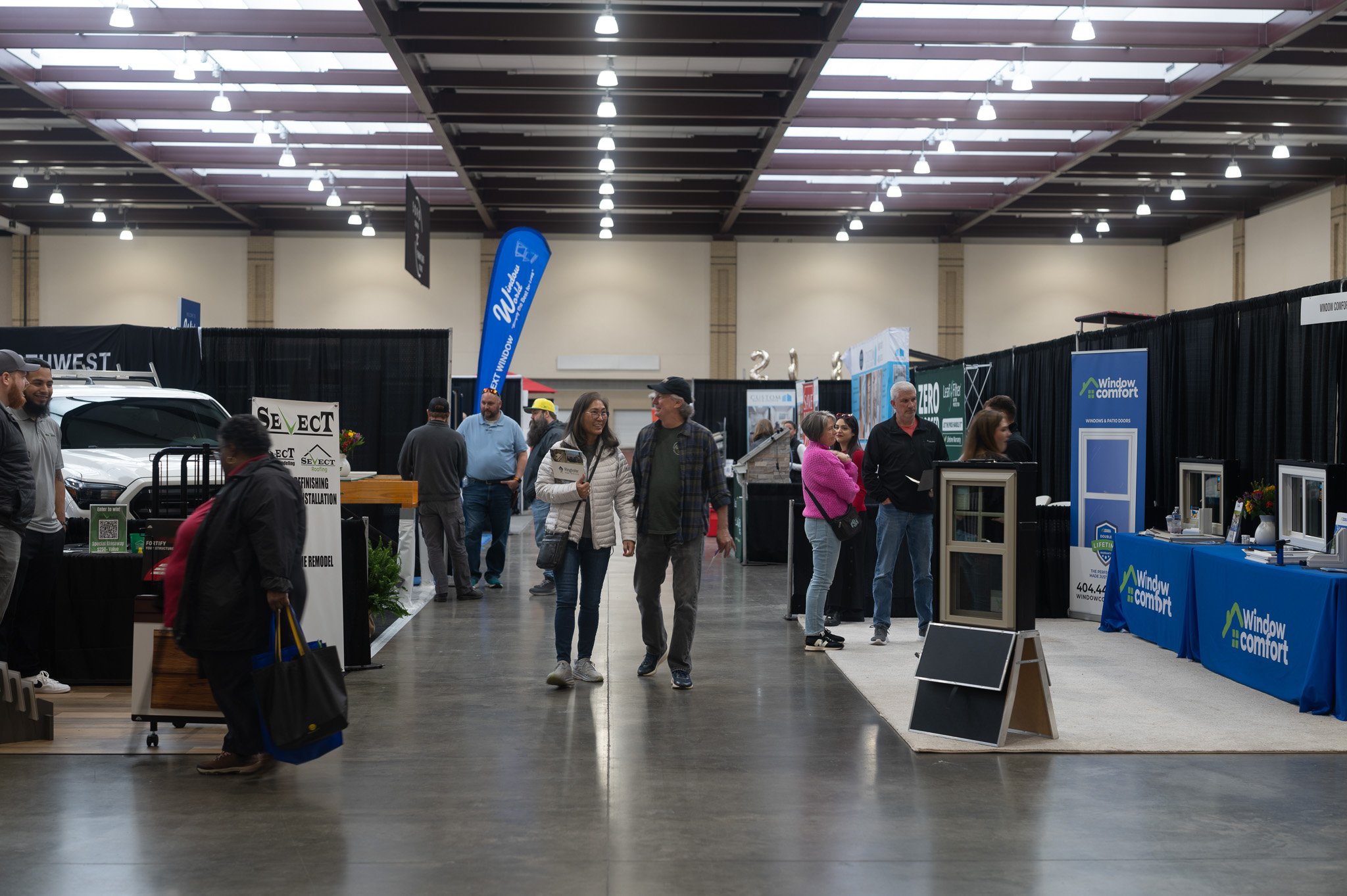 People walking and standing in an indoor trade show or exhibition hall, with booths and displays on the sides showcasing window and door products.