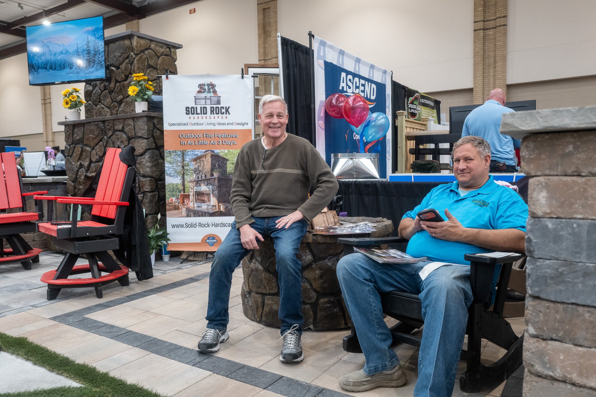 Two men sitting in front of a display booth at an indoor event or trade show. One is smiling and standing, while the other is sitting and looking at his phone. The booth features signage for Solid Rock Hardscapes and has outdoor fire features adverti