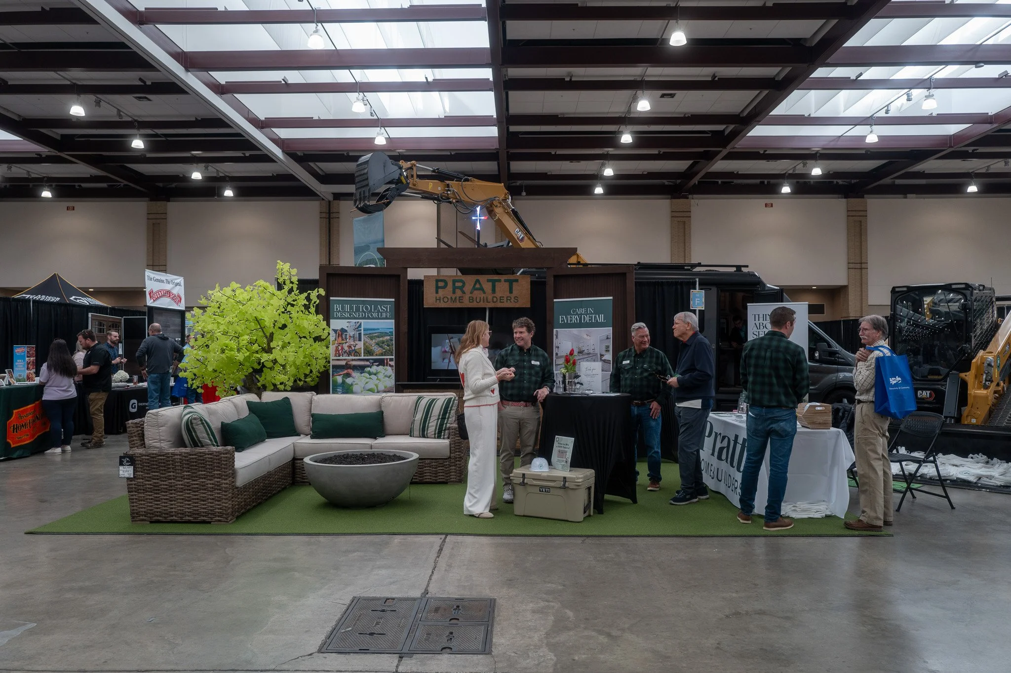An indoor exhibition with various booths and displays, including furniture and construction equipment, and a group of people conversing in front of a booth labeled 'Pratt Home Builders'.