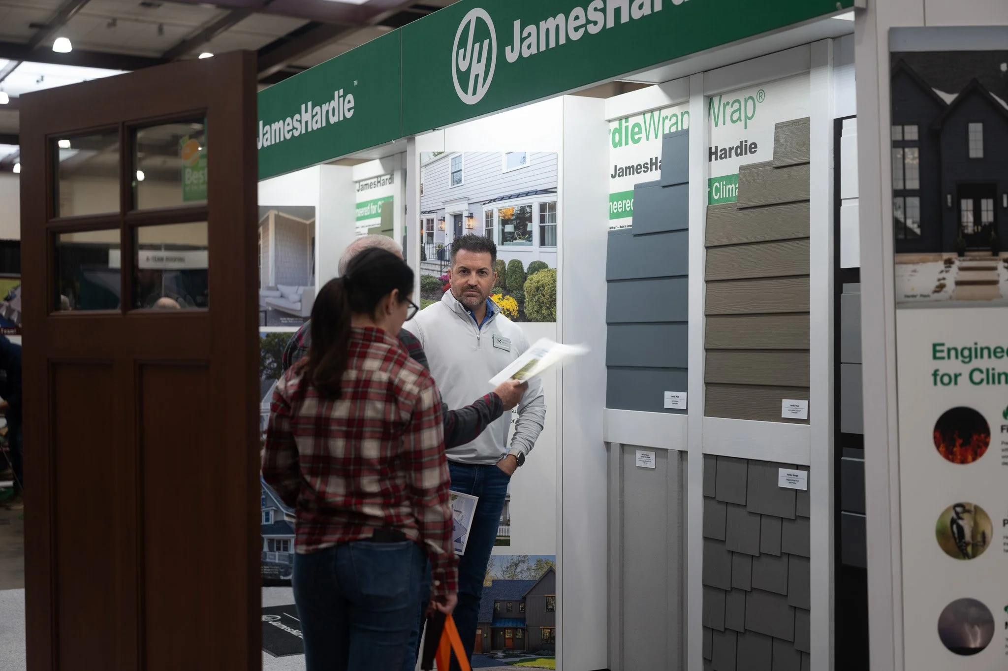 People browsing a home exterior display at a trade show, with siding and cladding samples, and a large sign reading 'JamesHardie' and 'JamesHardieWrap'.