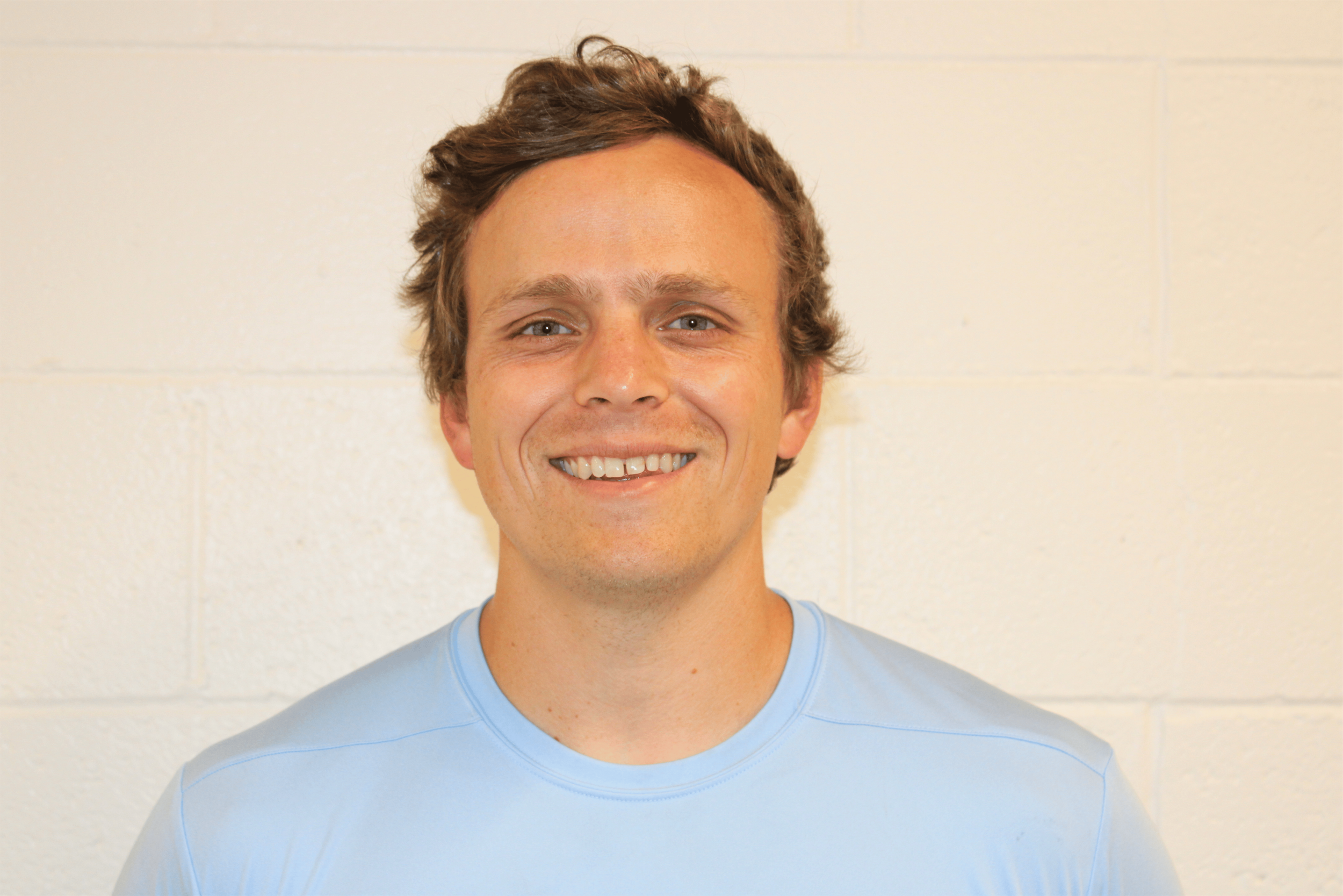 A young man with short, wavy brown hair and blue eyes, smiling, wearing a light blue T-shirt, standing against a plain, off-white wall.