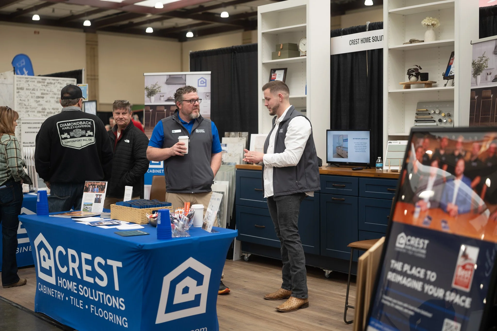 A booth at a home solutions exhibition featuring Crest Home Solutions. The booth has a blue tablecloth with the company logo and text listing services like cabinetry, tile, and flooring. Two representatives are talking to visitors, one holding a coff