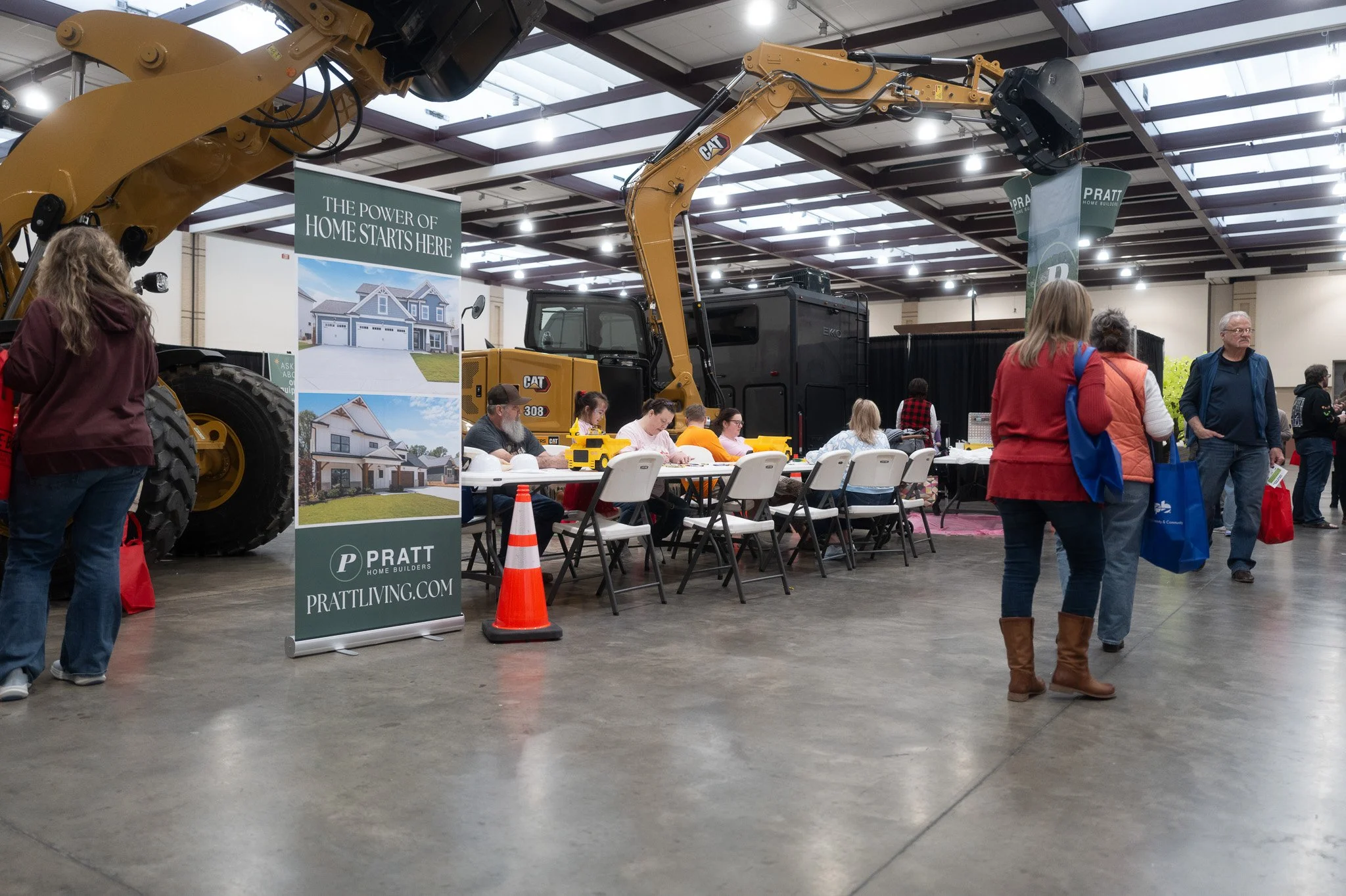 Indoor event showcasing construction equipment, including a large excavator, with people seated at tables and walking around, and promotional banners from Pratt Home Builders.
