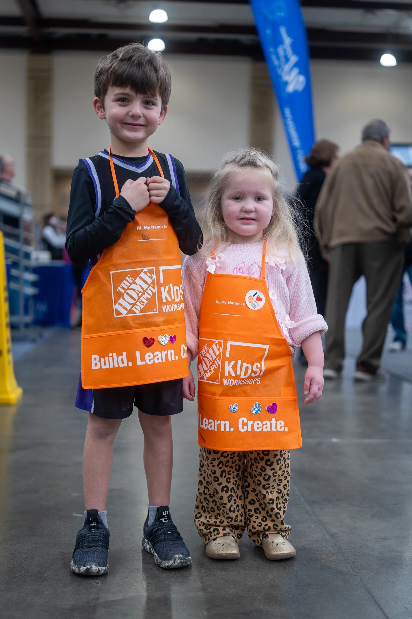 Two children standing side by side at an indoor event, both wearing orange Home Depot Kids Workshops aprons. The boy on the left is smiling, has short brown hair, and is wearing black athletic shorts and black sneakers. The girl on the right has blon