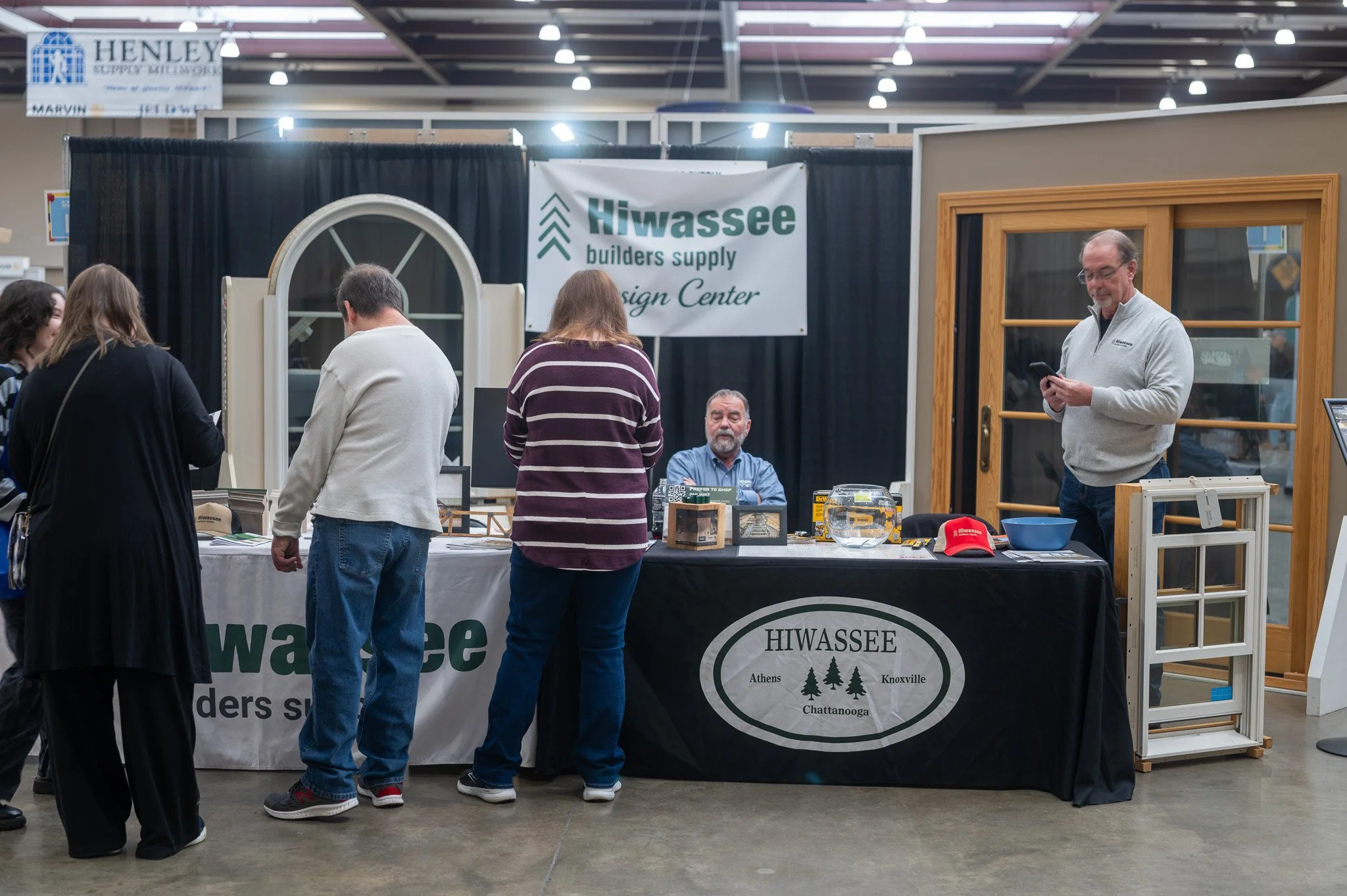 People browsing a booth for Hiwassee Builders Supply at an indoor event. The booth has a black tablecloth with promotional items, and a man in a gray sweater is looking at his phone. The backdrop features a sign with the Hiwassee Builders Supply logo