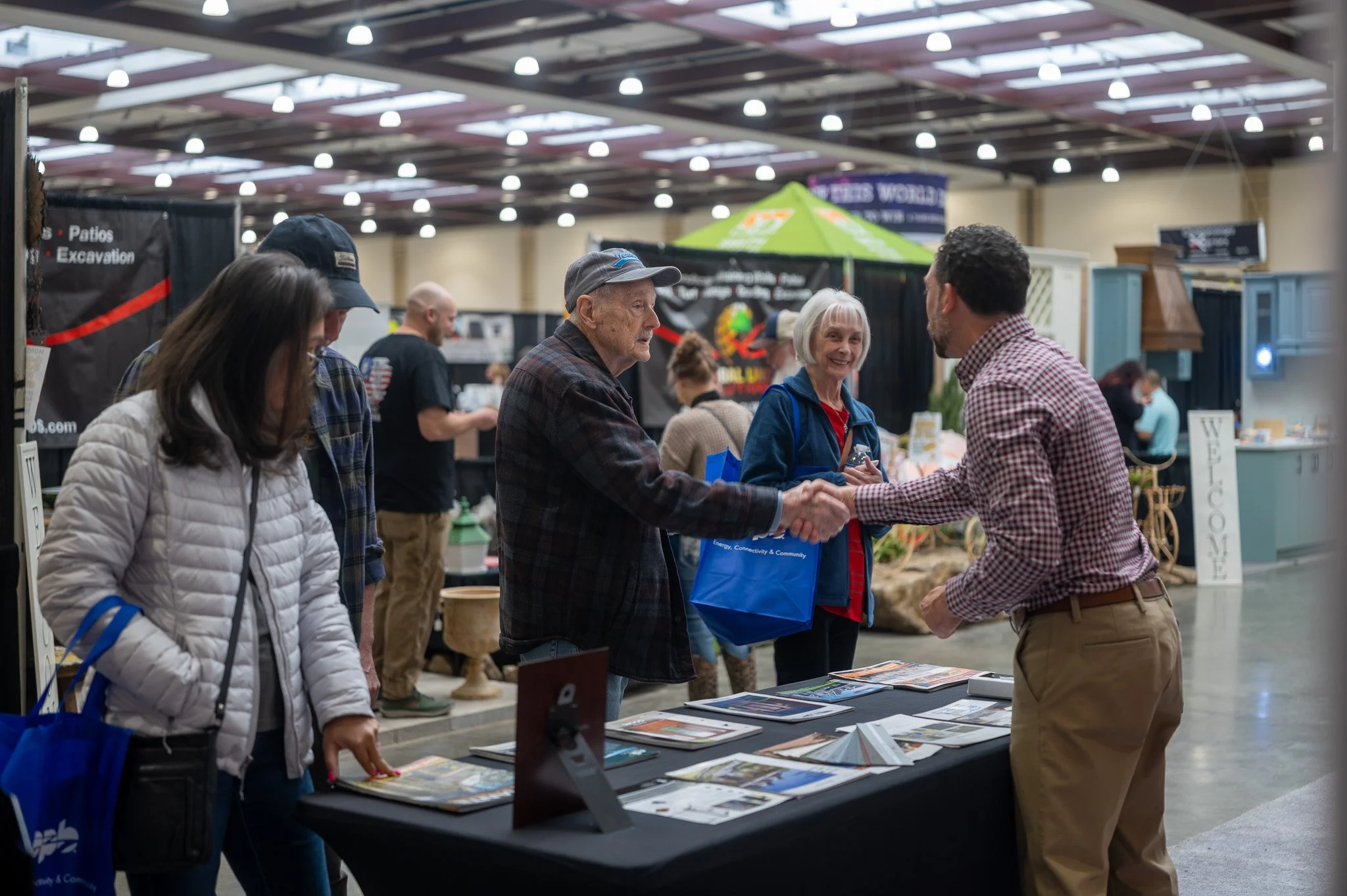People attending a conference or trade show, engaging in conversations around a booth with informational brochures and displays, inside a large indoor venue.