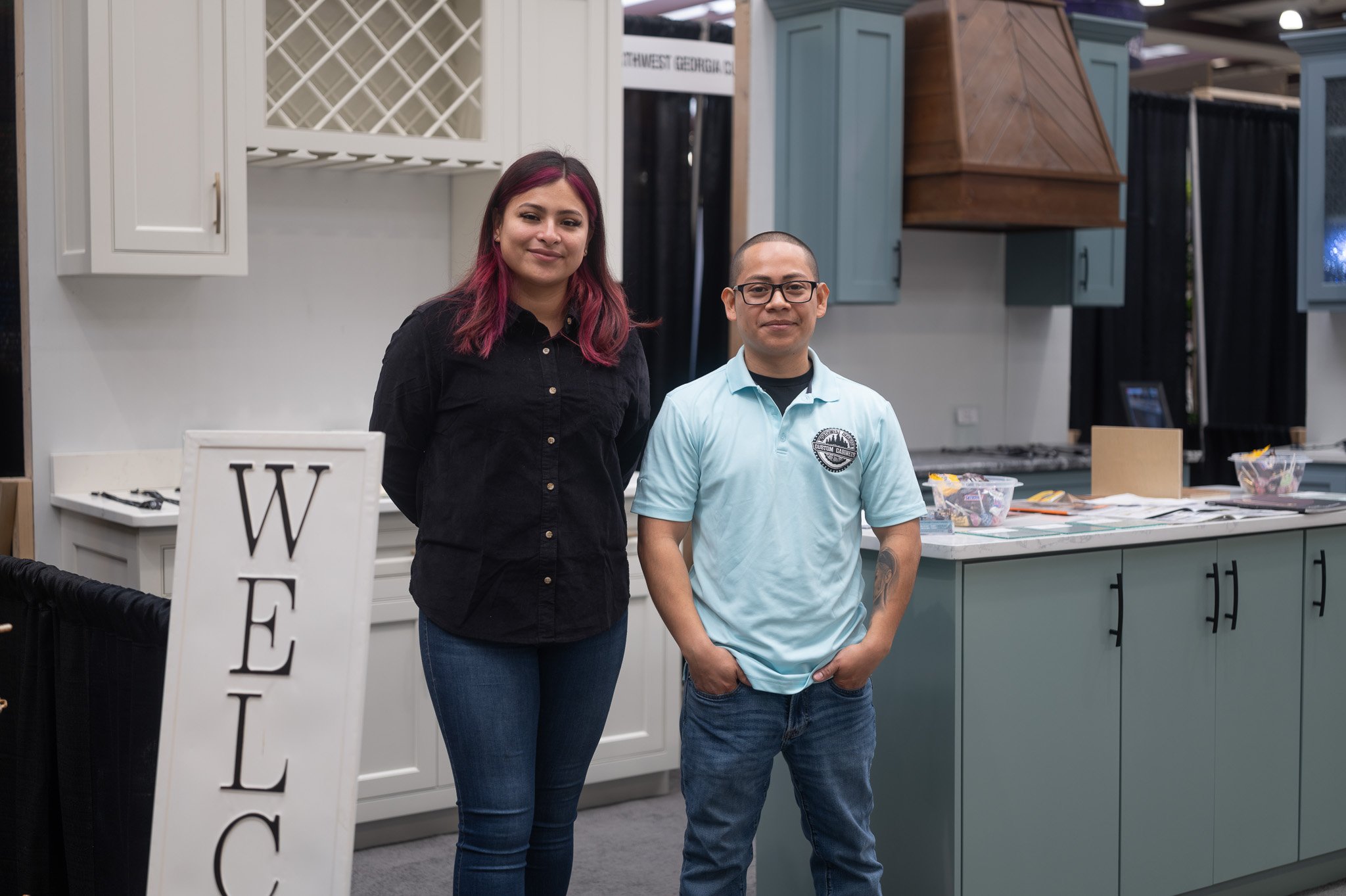A woman with long hair dyed pink and a man with glasses, both standing in front of a display booth with cabinetry units at an indoor event.