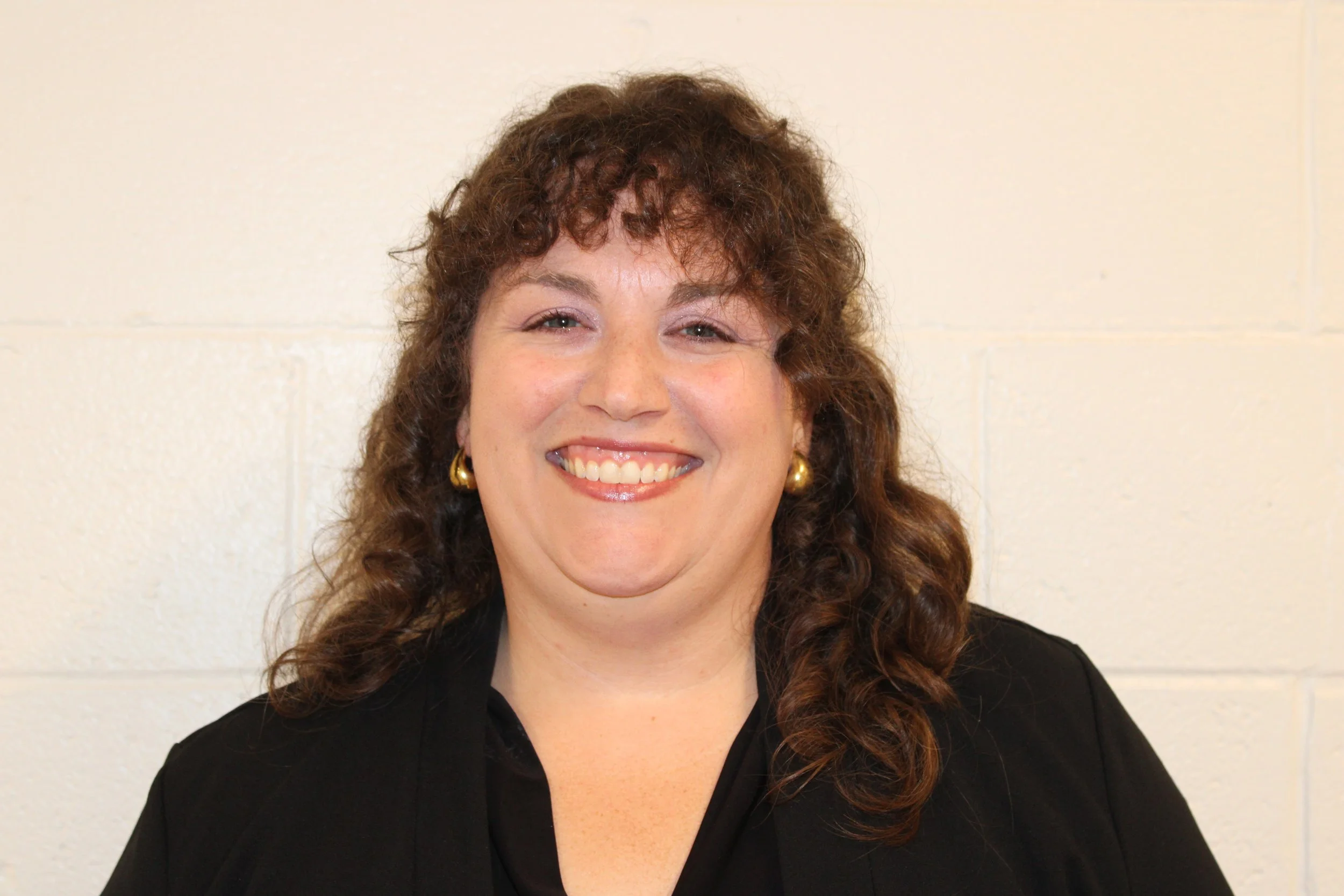 A smiling woman with curly brown hair, wearing gold earrings and a black top, standing against a plain light-colored wall.