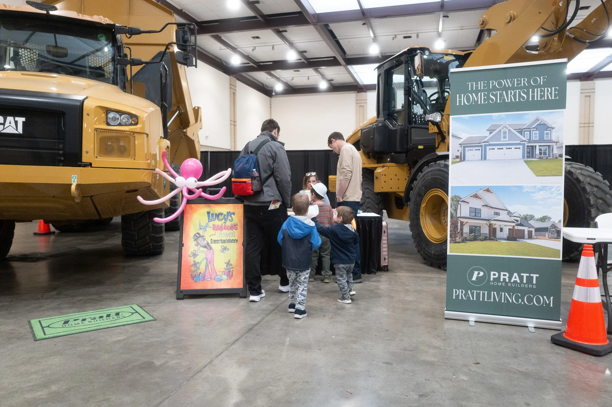 Children and adults gathered around construction equipment at an indoor home building expo, with a sign promoting homebuilding and balloons for entertainment.