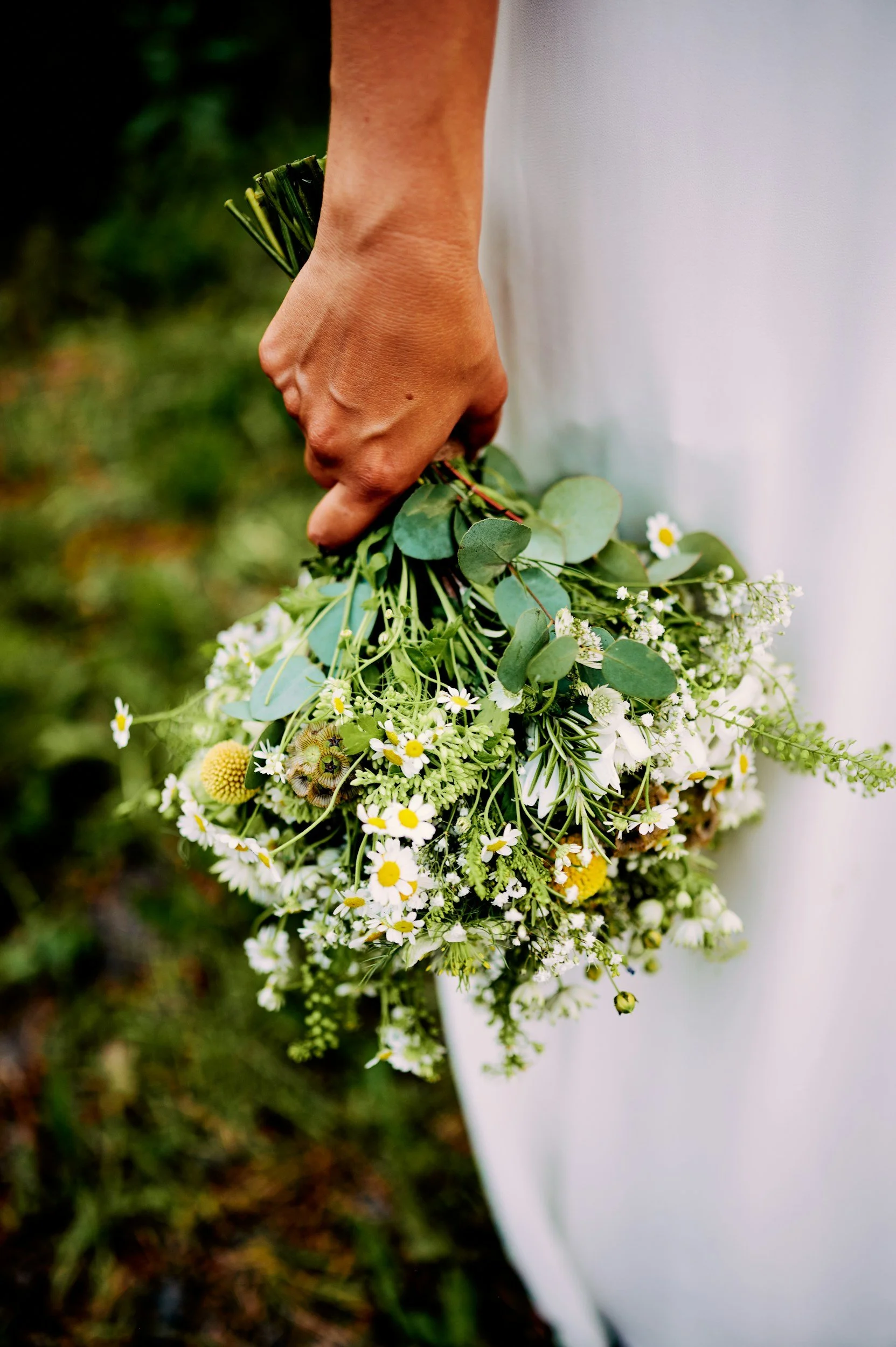 A person in a white garment holding a bouquet of white and yellow flowers and greenery outdoors.