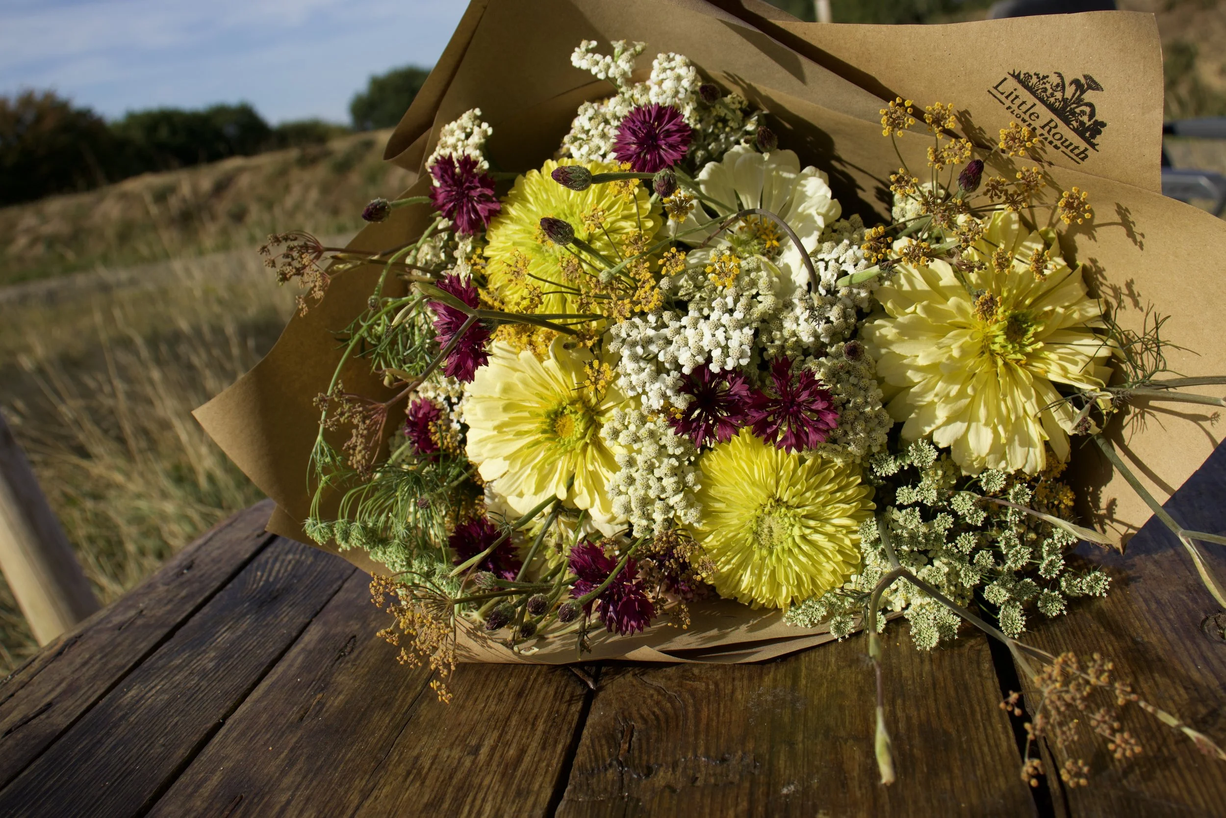 Bouquet of mixed flowers including yellow, purple, and white blooms wrapped in brown paper, resting on a rustic wooden surface outdoors.