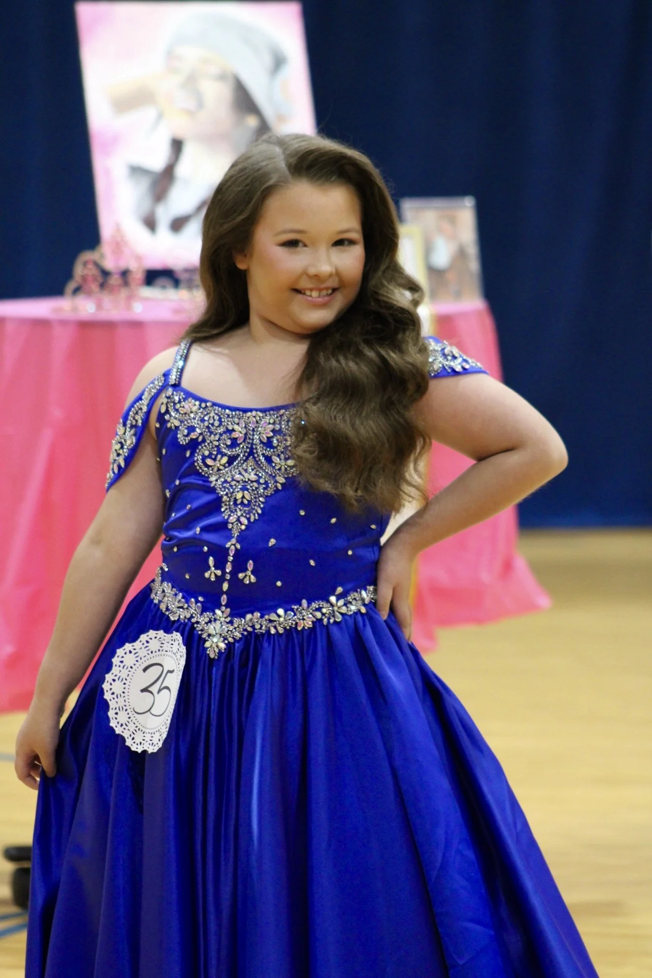 Young girl in a royal blue dress with silver embroidery, posing with a smile at a dress competition, wearing a badge with number 35, in a decorated indoor setting with pink drapes and framed photographs in the background.