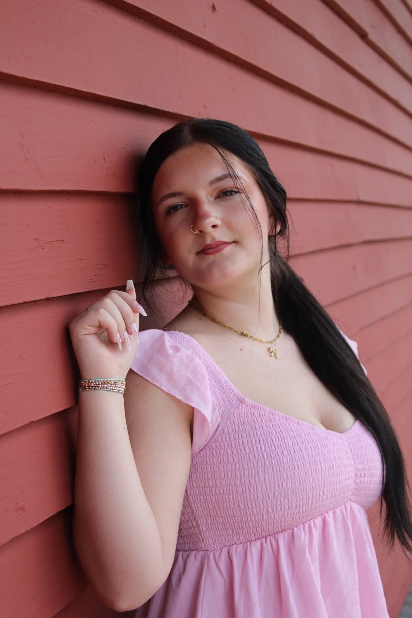 A young woman with dark hair and light skin leaning against a pink wooden wall, wearing a pink dress and multiple bracelets.