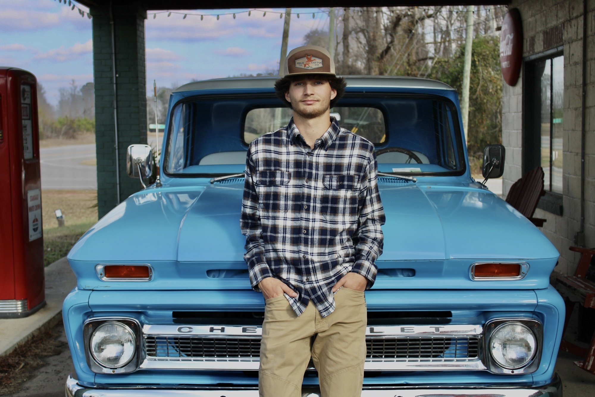 A young man with dark, curly hair wearing a beige hat, plaid shirt, and khaki pants, leaning against the front of a vintage blue Chevrolet truck at a retro gas station.