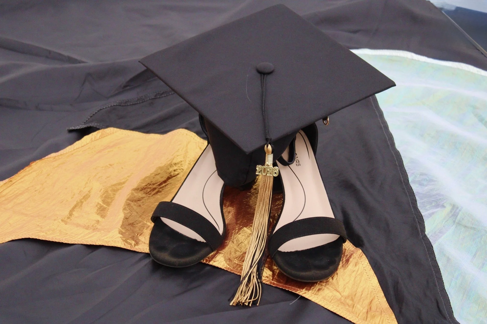 Graduation cap, diploma, and a gold tassel with the year 2026 hanging from the cap, placed on a black and gold fabric background.