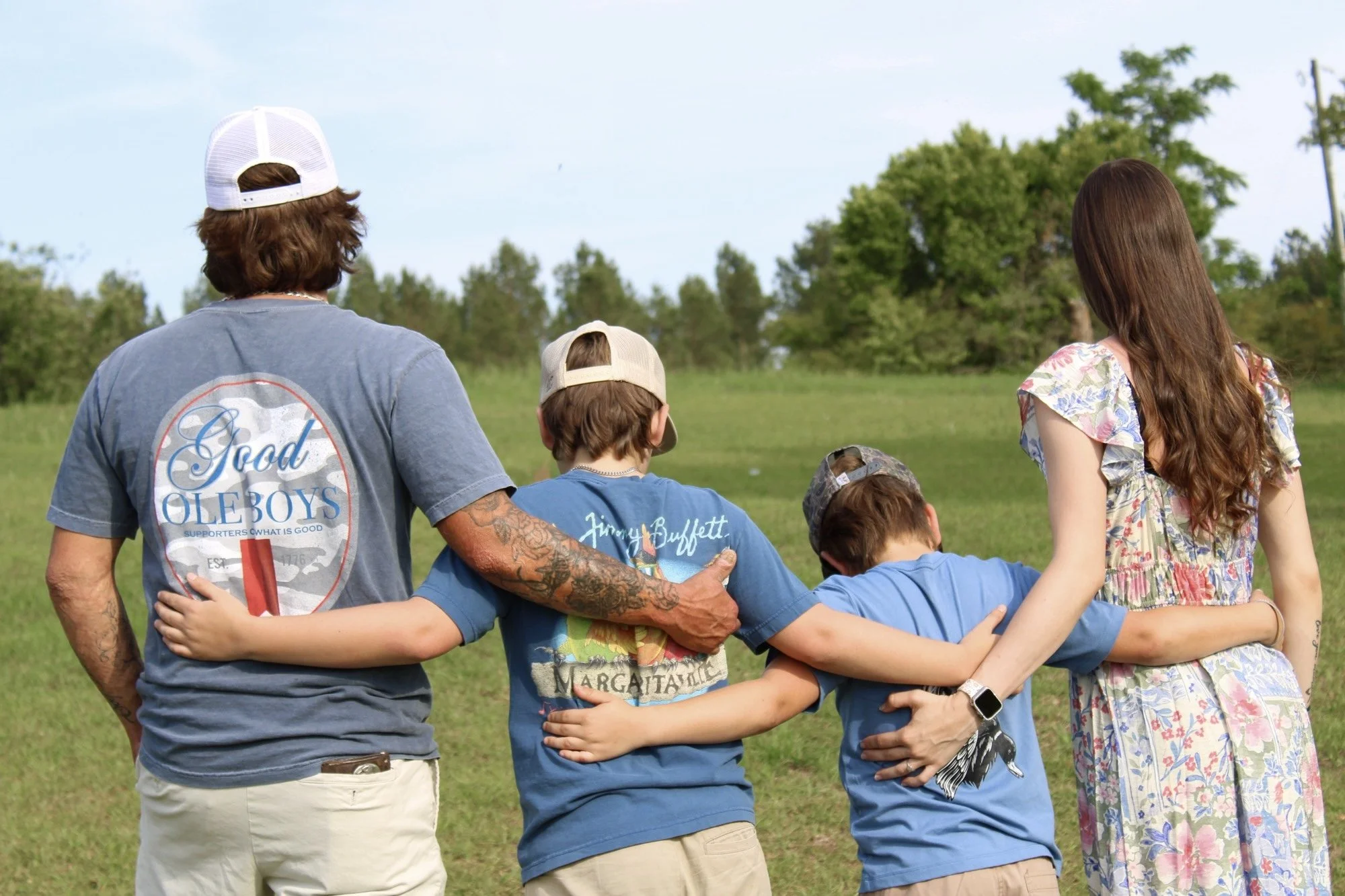 A group of people standing in a circle outdoors, with their arms around each other's shoulders. The group includes a man with a tattooed arm and a woman with long brown hair, both facing away from the camera, and children wearing blue shirts. They are on a grassy field with trees and a cloudy sky in the background.