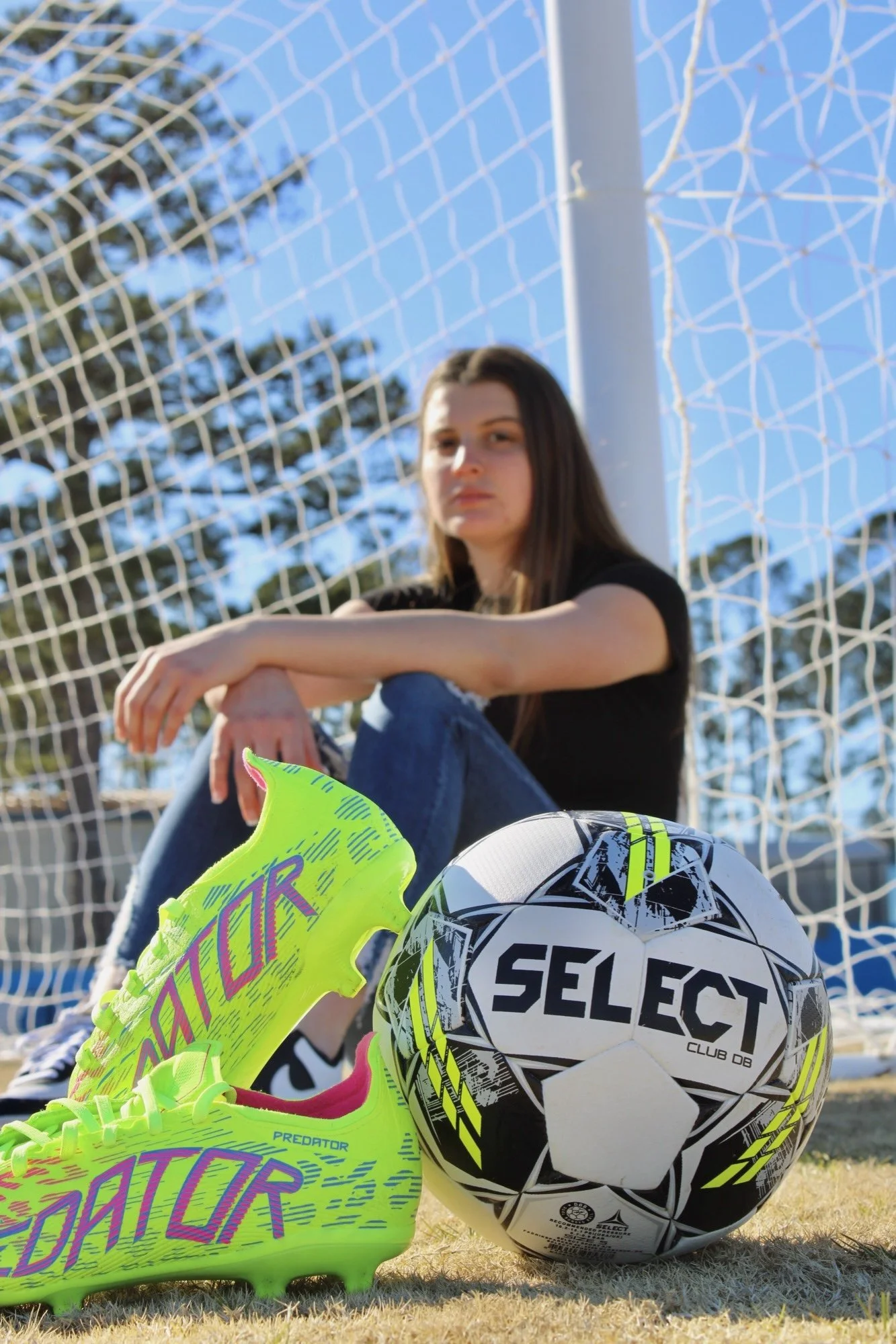 A young woman with long dark hair sitting on a soccer field near the goalpost, with soccer balls and colorful cleats in the foreground and a bright blue sky in the background.