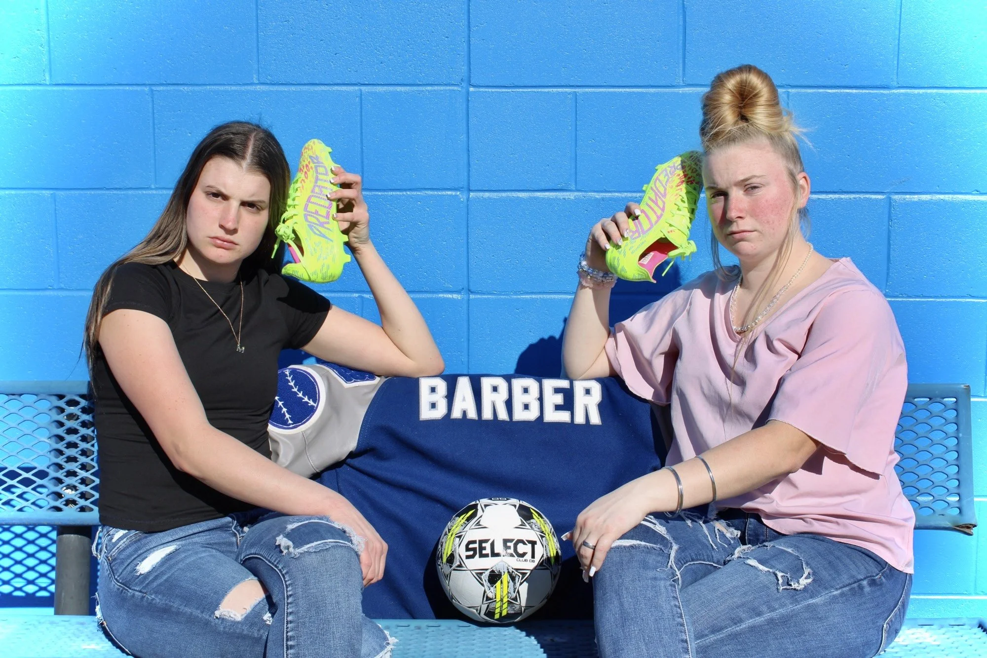 Two young women sitting on a blue bench against a blue wall, holding bright yellow soccer cleats to their heads, with a soccer ball between them.