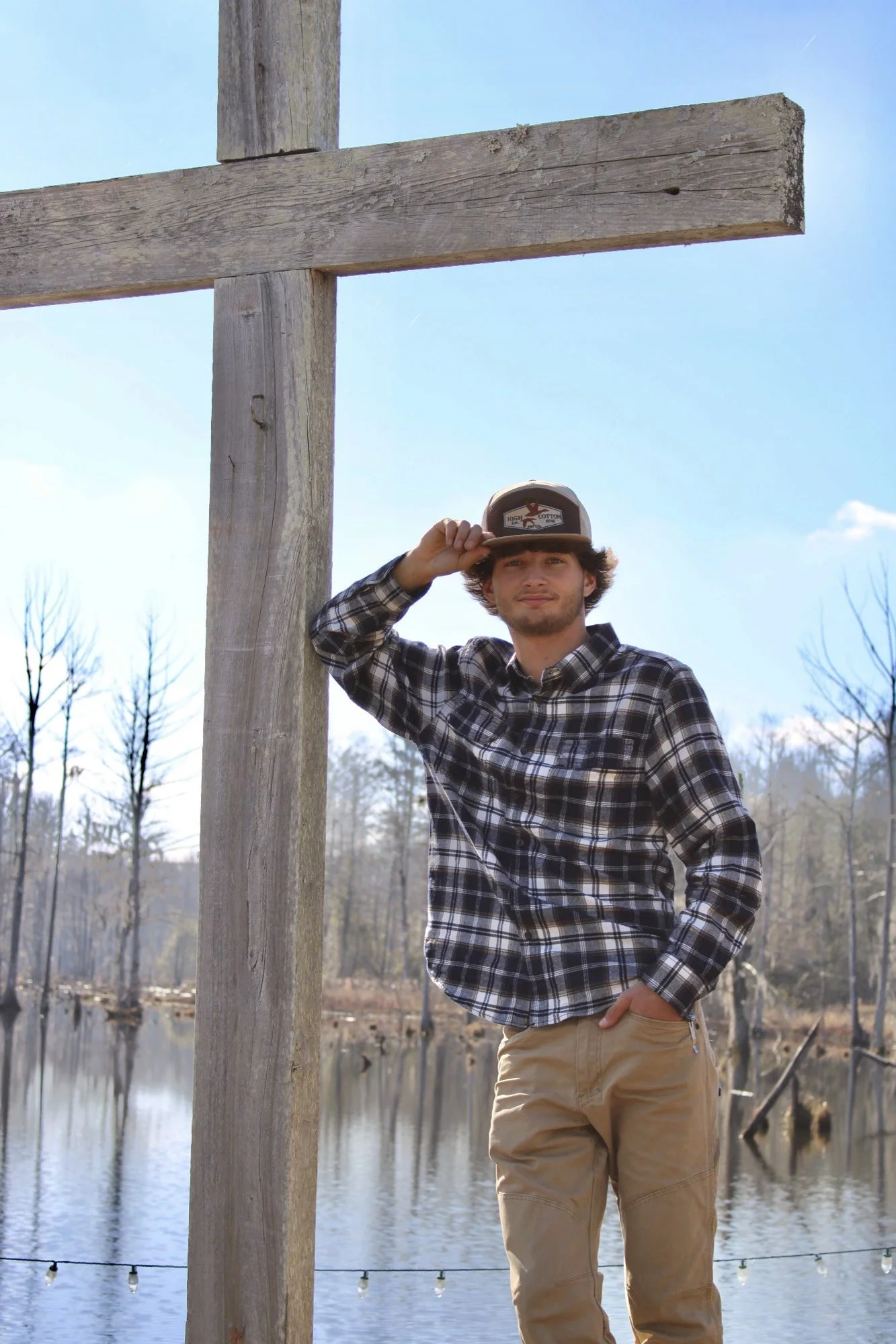 A young man wearing a plaid shirt, khaki pants, and a trucker hat standing outdoors by a body of water with a wooden cross and string lights in the background.