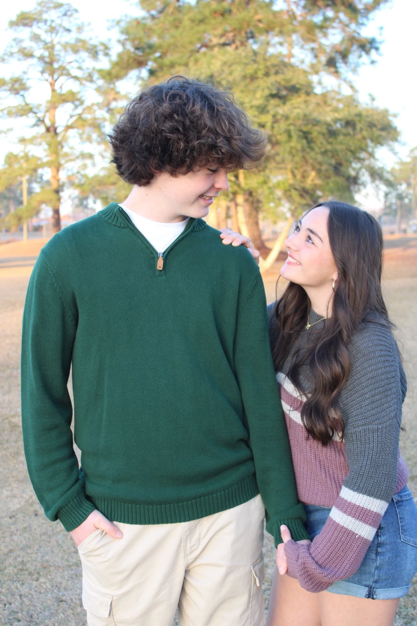 A young man with curly hair smiling and looking at a young woman with long dark hair who is smiling back and touching his shoulder in an outdoor park setting with trees in the background.