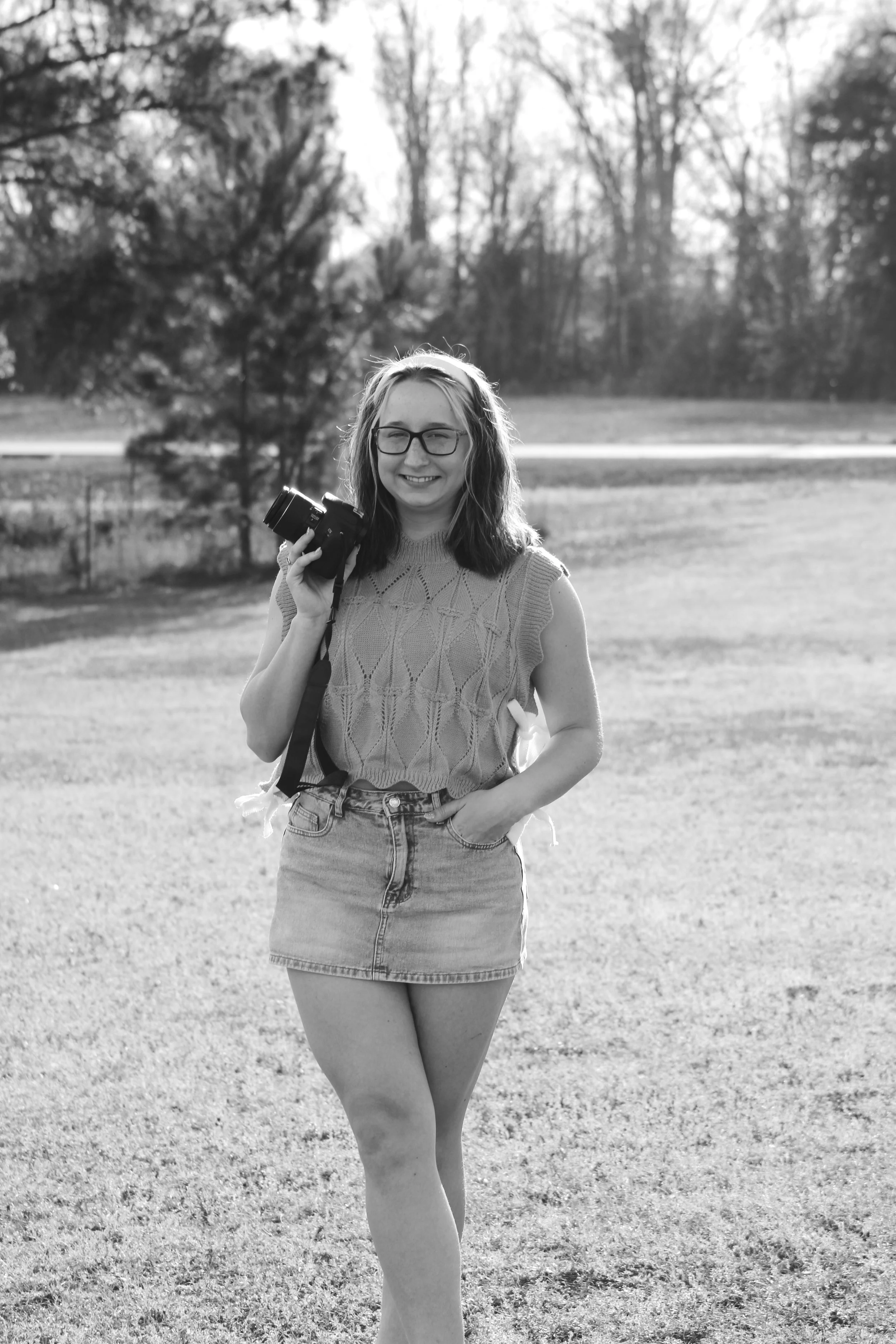 A woman standing outdoors in front of trees, holding a camera, smiling, wearing glasses, a sleeveless knitted top, and a denim skirt.