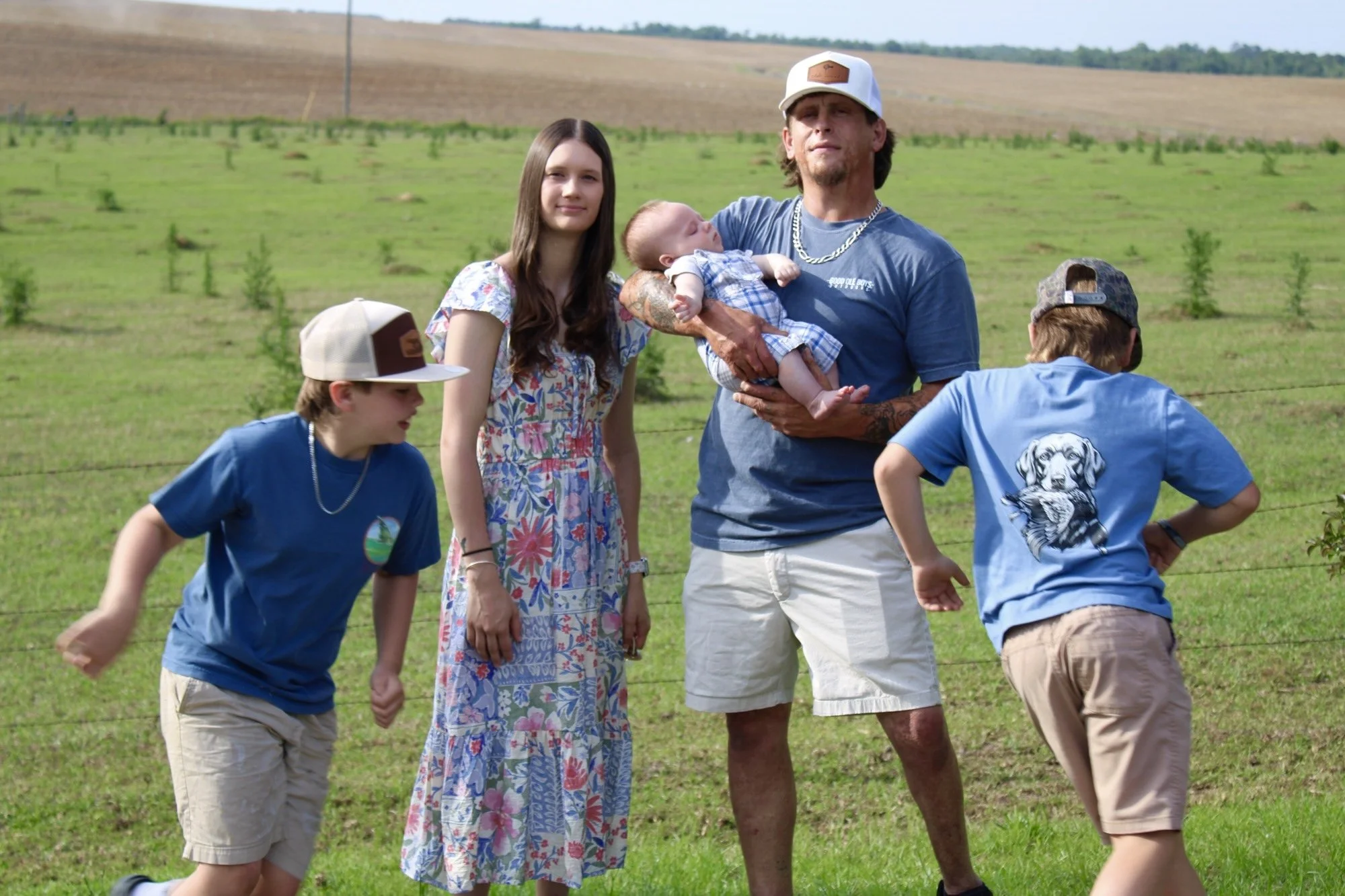 A group of people walking outdoors in a grassy field with hills in the background. A man is holding a baby, and a woman and two boys are nearby, one wearing a cap and the other with a baseball cap backward.