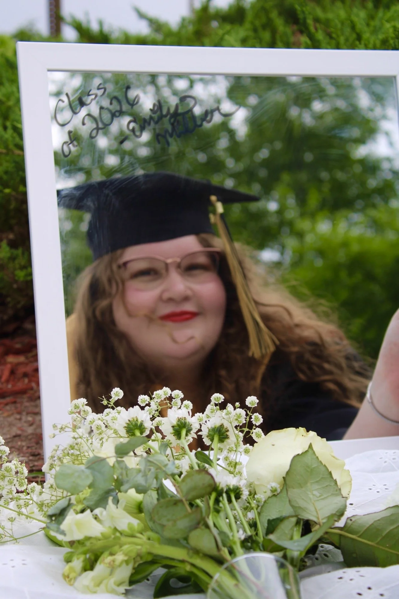 A woman in a graduation cap and gown takes a selfie with a mirror, with white flowers in front of her. The background shows greenery and an outdoor setting.