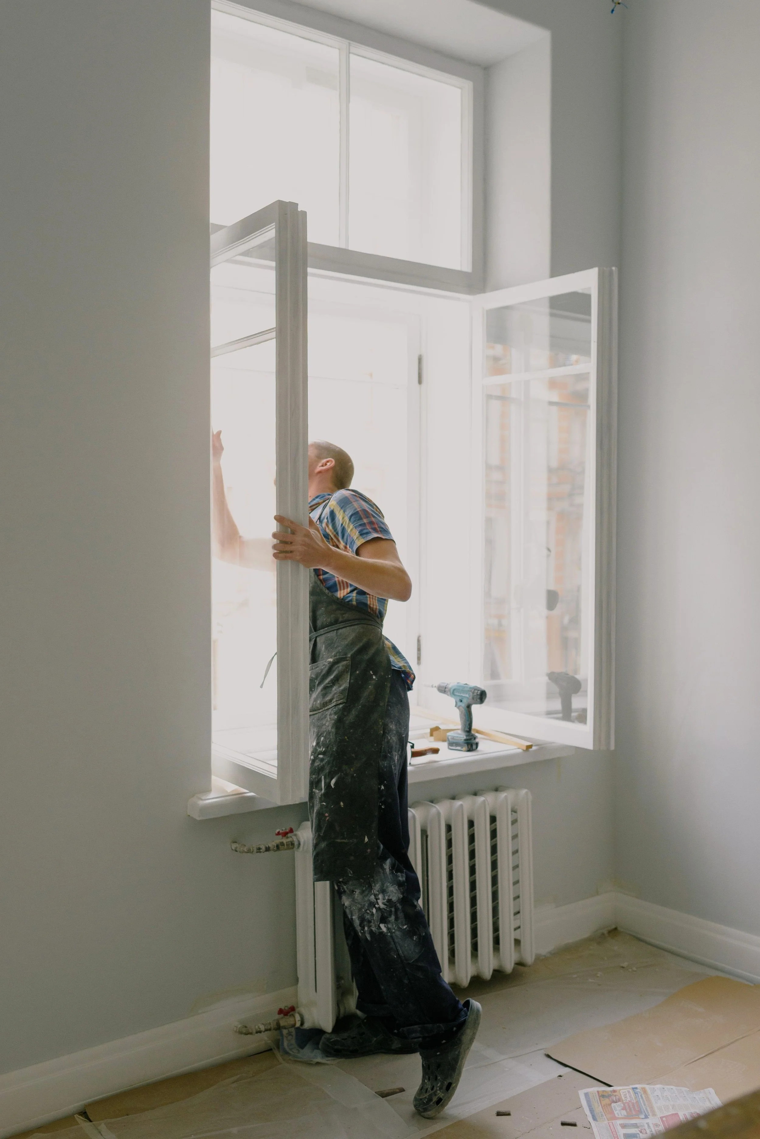 Man painting window frames white inside a room, with tools on the windowsill, a newspaper on the floor, and a radiator beneath the window.