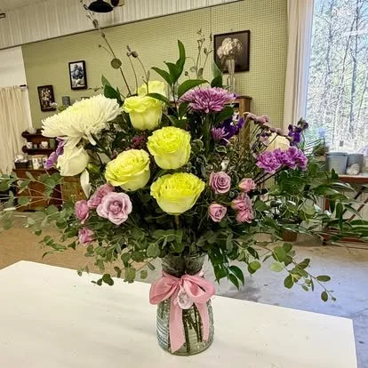 A colorful bouquet of flowers in a glass jar with a pink ribbon, placed on a white surface in a cozy room with framed pictures and large windows.