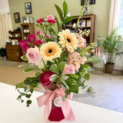 A floral arrangement in a pink vase with pink ribbon, containing pink roses, yellow gerbera daisies, and assorted greenery, on a white surface in a room with wooden furniture and plants.