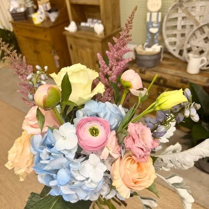 Colorful bouquet of flowers including roses, peonies, and other blooms in a store setting with wooden furniture and decorative items in the background.