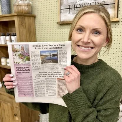 A woman with blonde hair and a green sweater smiling and holding up a newspaper indoors, with shelves and a wooden sign in the background.