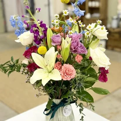 A colorful bouquet of mixed flowers including lilies, roses, daisies, and other blooms in a light green vase with a dark green ribbon, placed on a table indoors.