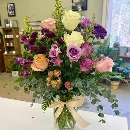 A colorful bouquet of roses, lisianthus, and other flowers in a glass vase with a cream ribbon, set on a table indoors.