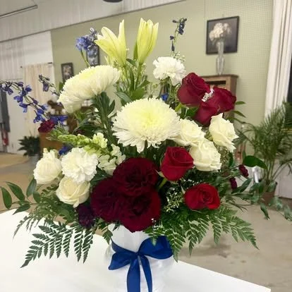 A bouquet of mixed flowers including white lilies, white carnations, red roses, purple flowers, and green foliage, arranged in a blue ribbon-tied vase.