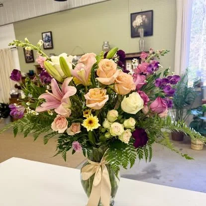 A large bouquet of mixed flowers in a glass vase with a cream-colored ribbon, featuring pink lilies, peach roses, white roses, purple statice, yellow daisy-like flowers, and various greenery, on a white surface in a room with green walls and framed p