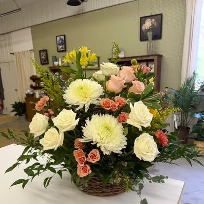 A large floral arrangement in a wicker basket featuring white roses, yellow lilies, pink roses, white chrysanthemums, and other greenery in an indoor setting.