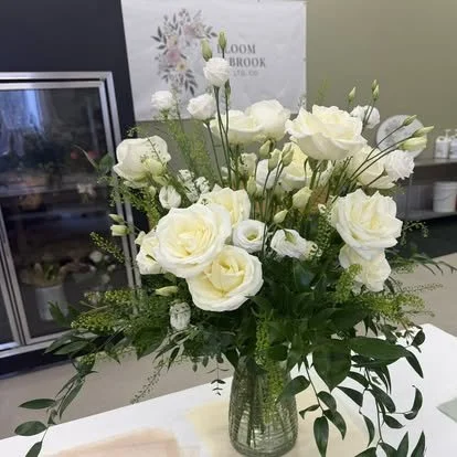 A bouquet of white roses and other white flowers in a glass vase, with green leaves and delicate stems.