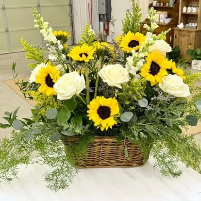 A floral arrangement in a wicker basket containing white roses, yellow sunflowers, and various green foliage.
