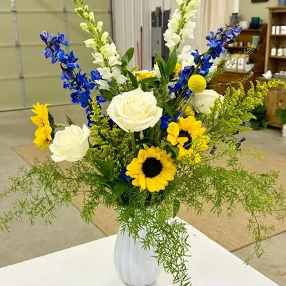 Fresh flower bouquet with white roses, yellow sunflowers, blue delphiniums, white snapdragons, and greenery in a white vase.