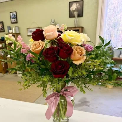 A bouquet of multicolored roses and flowers in a glass vase with a pink ribbon, placed on a table in a room with framed pictures on the wall and a window in the background.
