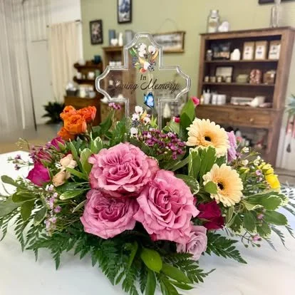 Floral arrangement with pink roses, yellow gerbera daisies, orange roses, and purple flowers in a room with shelves and framed pictures. Includes a memorial cross with the words "In Loving Memory."