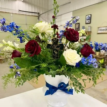 A floral arrangement in a white vase with a blue ribbon, containing red, white, and blue flowers with green foliage, set indoors on a table.