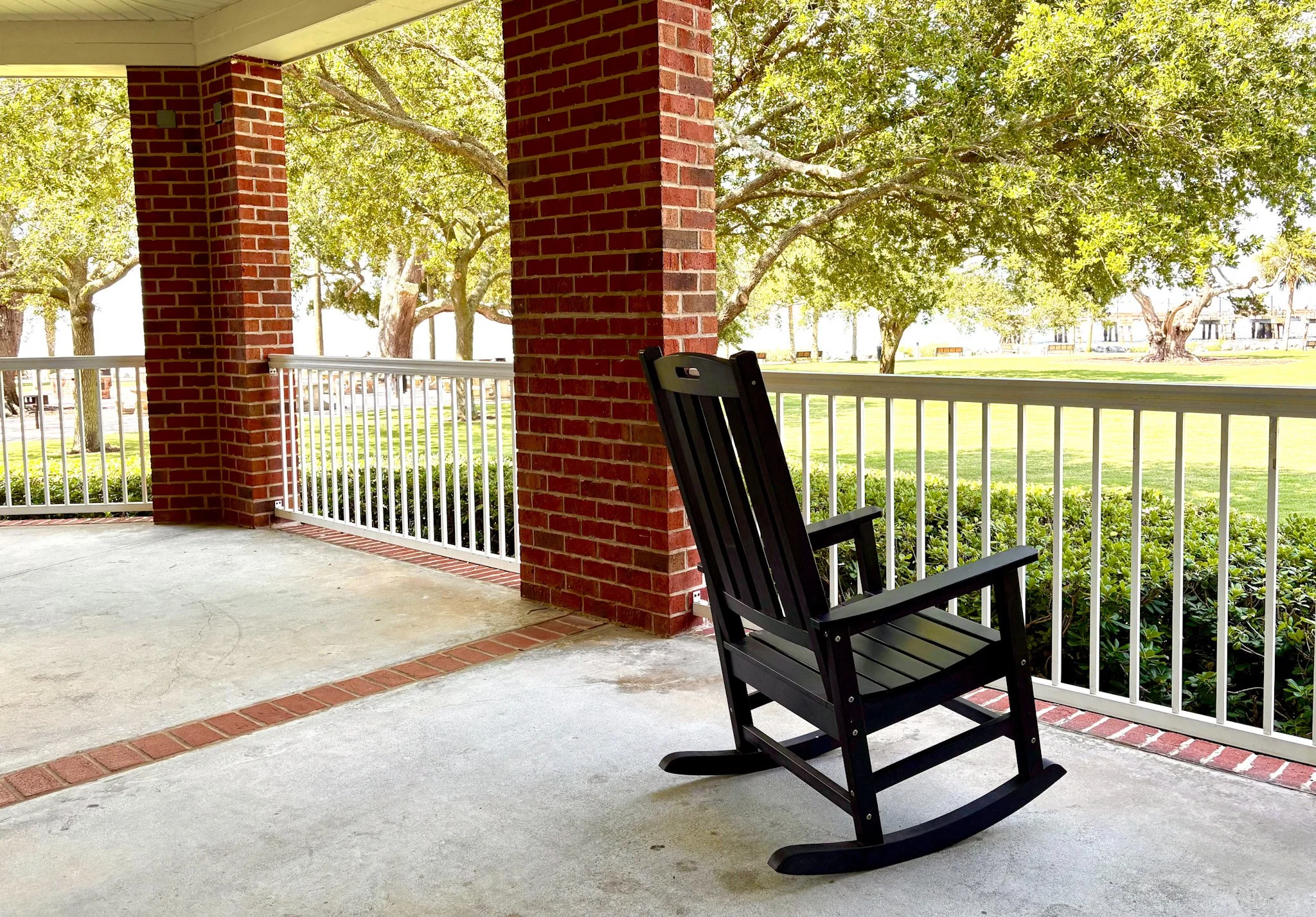 A rocking chair on a concrete and brick patio with brick pillars. A view beyond the patio of live oak trees and the ocean in the distance
