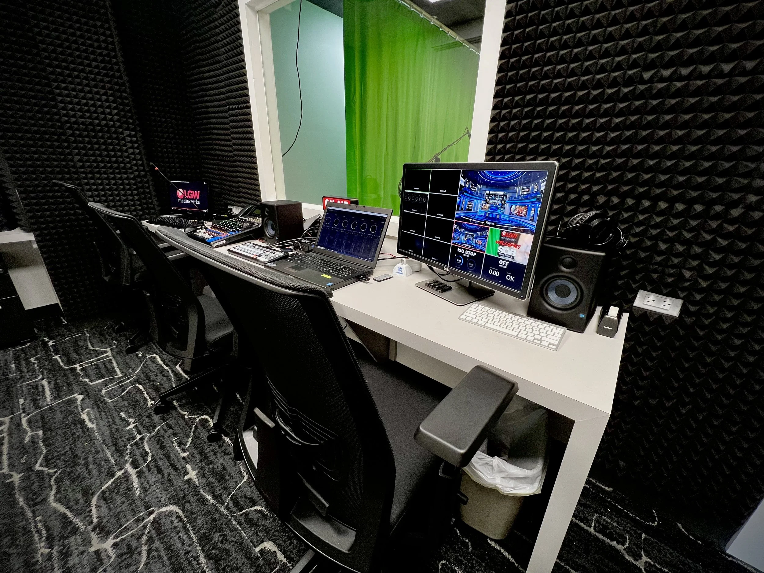 A recording studio with soundproof black foam walls, a white desk with multiple electronic devices including a computer, monitors, speakers, and audio equipment. There's a laptop, a large monitor displaying a graphic interface, and smaller equipment like a mixer and headphones. A window with green and blue backgrounds can be seen in the room.