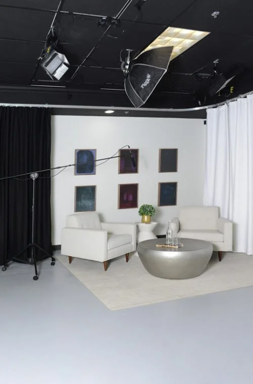 A photography studio with white and black curtains, two white armchairs, a round metallic coffee table with a glass object on it, a small green plant on a table, six framed artworks on the wall, lighting and camera equipment overhead.