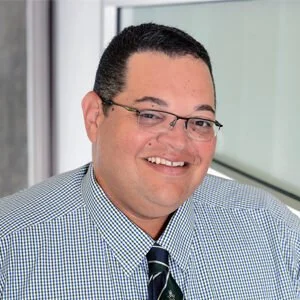 A man with glasses smiling, wearing a checkered shirt and a tie, standing by a window.